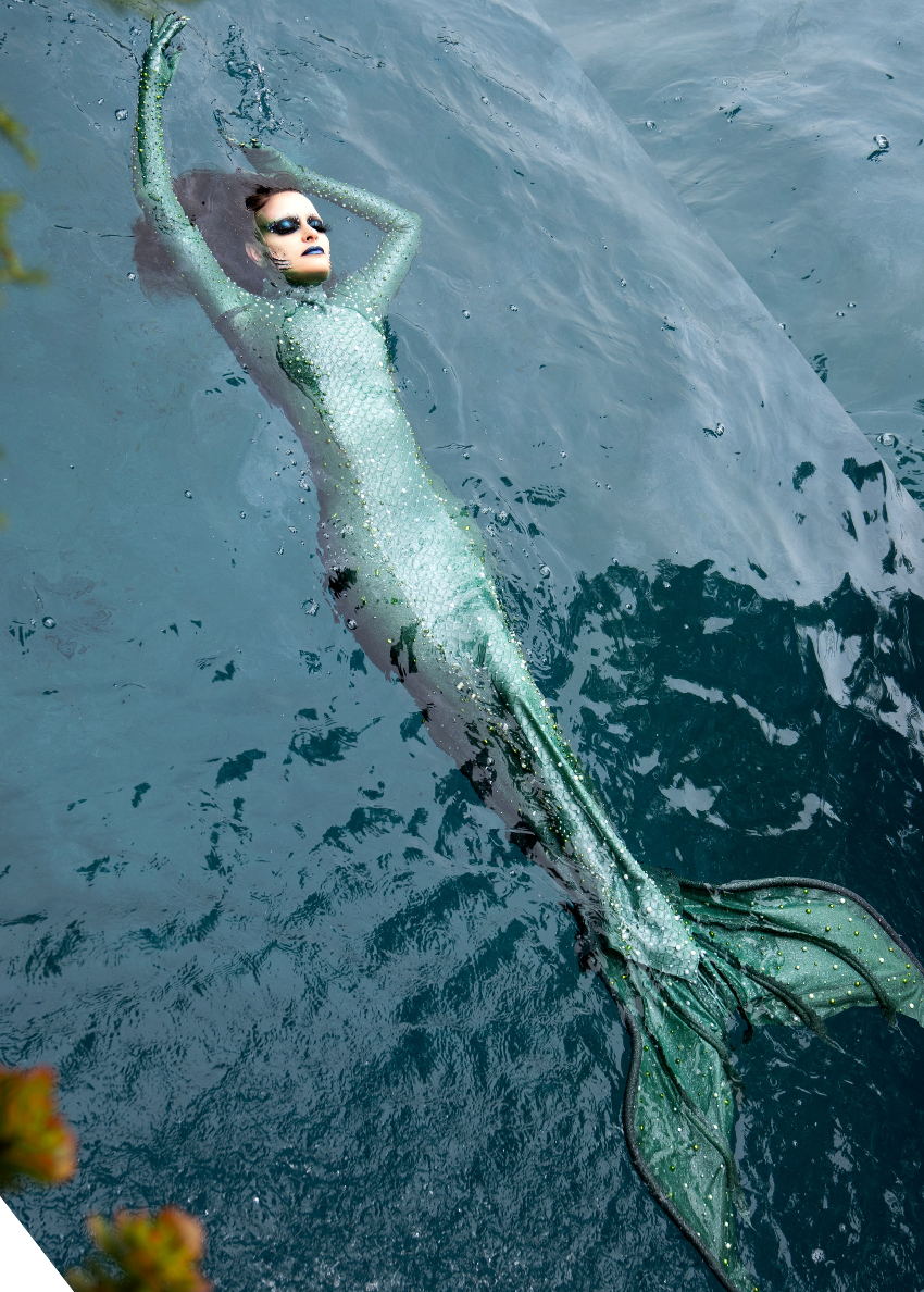 A person dressed as a mermaid in a sparkling, green, full-body costume floats on their back in clear, blue water, with arms raised and eyes closed.