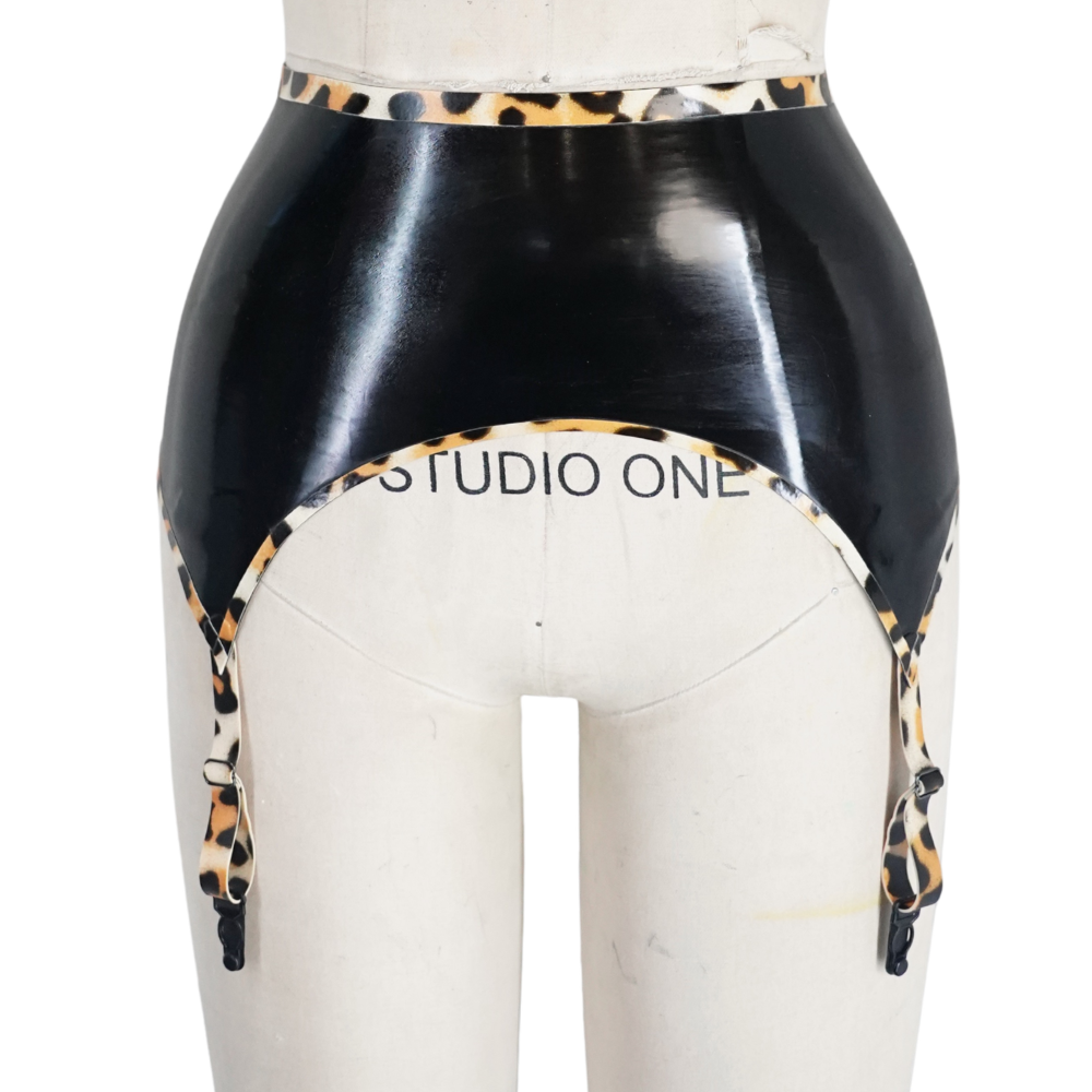 A black latex garter belt with leopard print trim by Vex Clothing, INC is shown on a white mannequin torso marked "STUDIO ONE," highlighted by a bold banner above to announce it's ready to ship.
