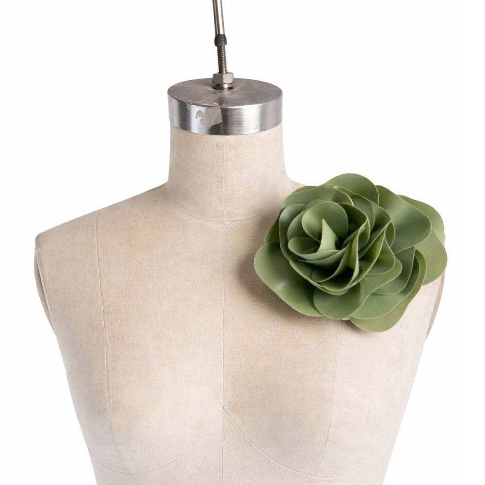 A beige dress form showcases a large, green, hand-crafted Vex Latex floral pin on its left shoulder. The stylized succulent- or camellia-inspired design makes it a bold statement accessory.