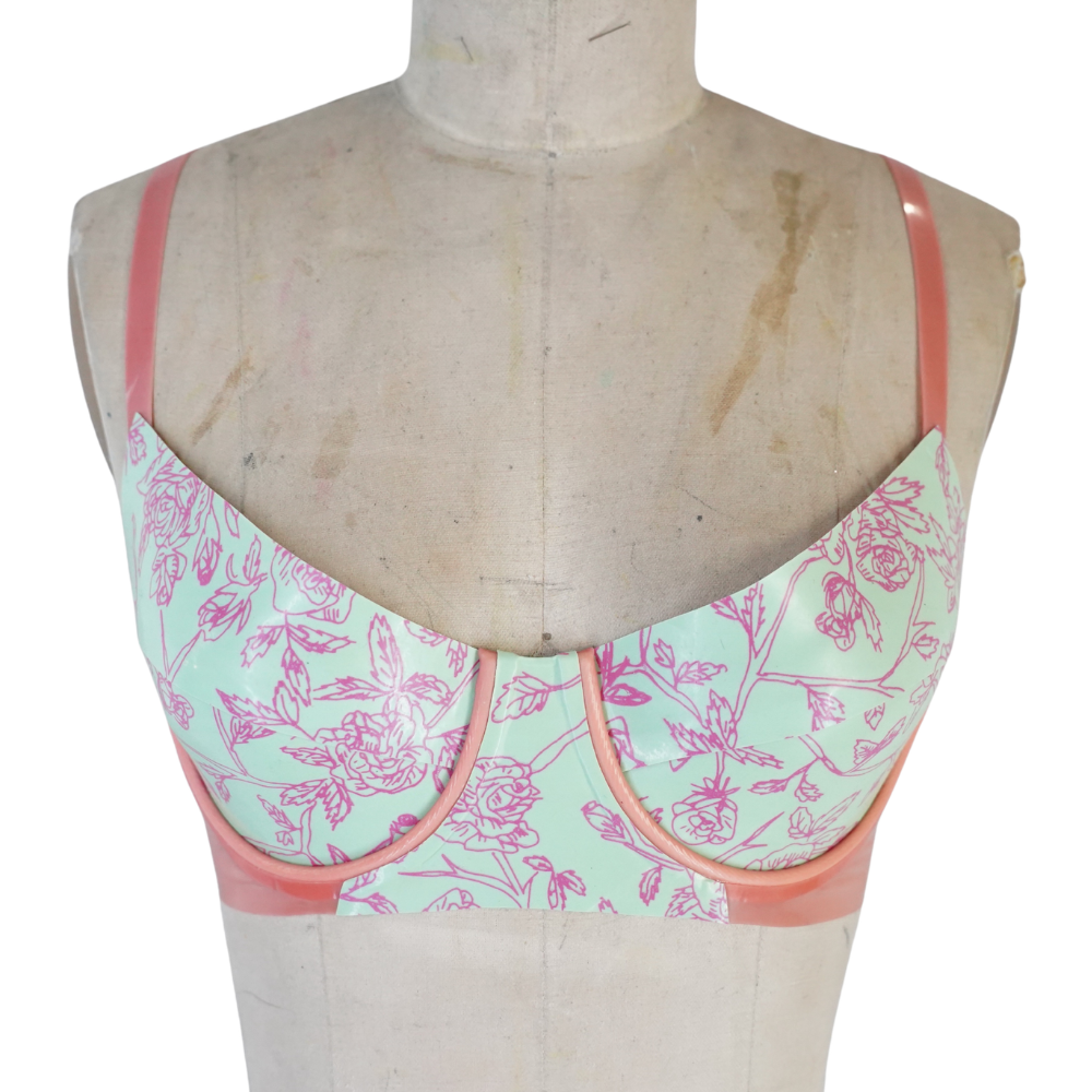 A dress form displays a Vex Inc. | Latex Clothing pastel bra with pink floral patterns, shiny peach straps, underwire support, and a mint green background. Ready to ship.