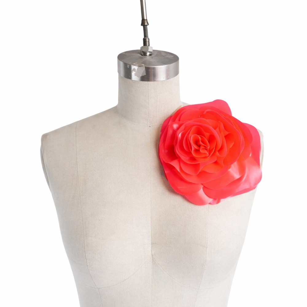 A Vex Latex dress form features a bold, sculptural flower brooch on the left shoulder, set against a plain white background.