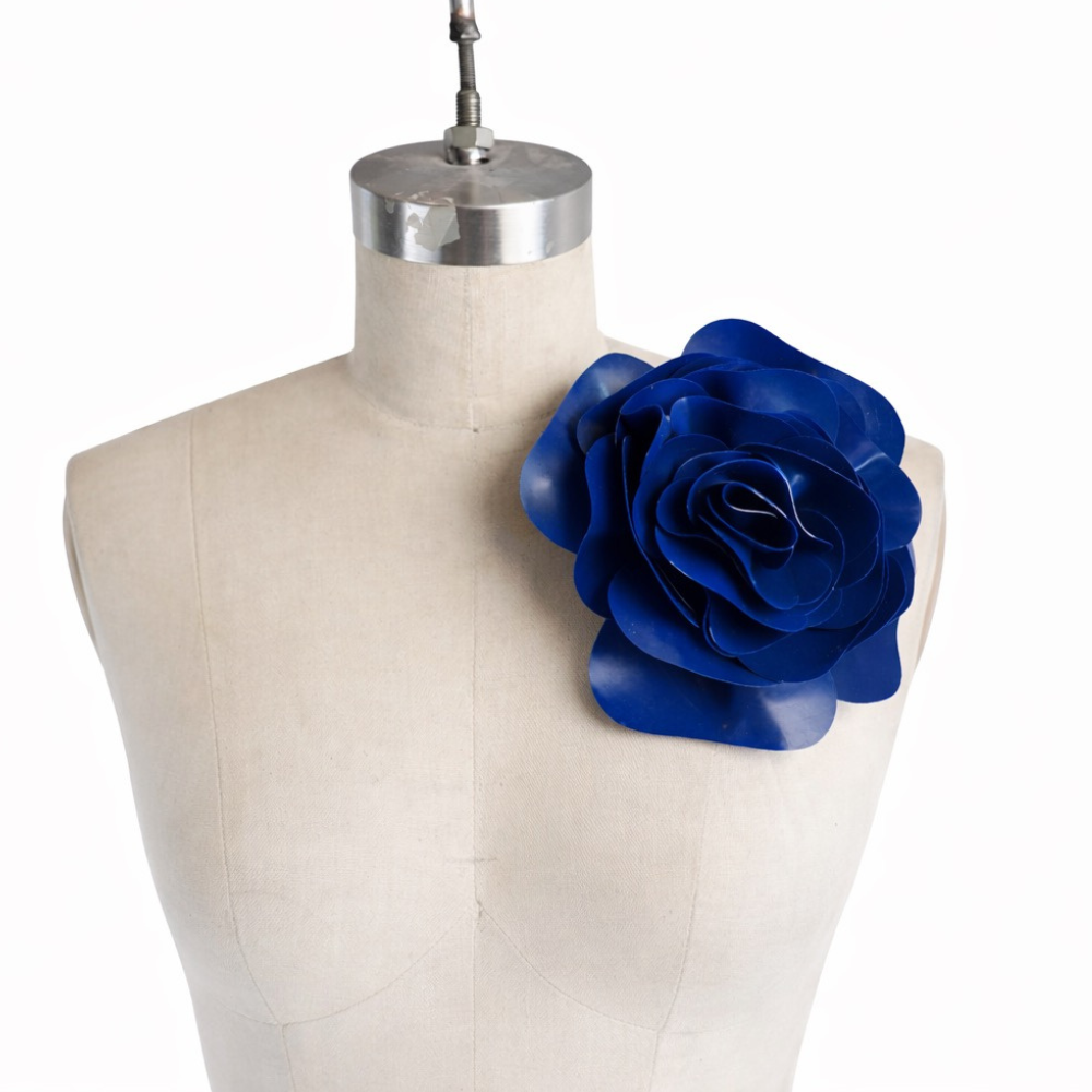 A Vex Latex dress form features an oversized, hand-crafted latex flower pinned to the left shoulder with a statement safety pin brooch, set against a plain white background.