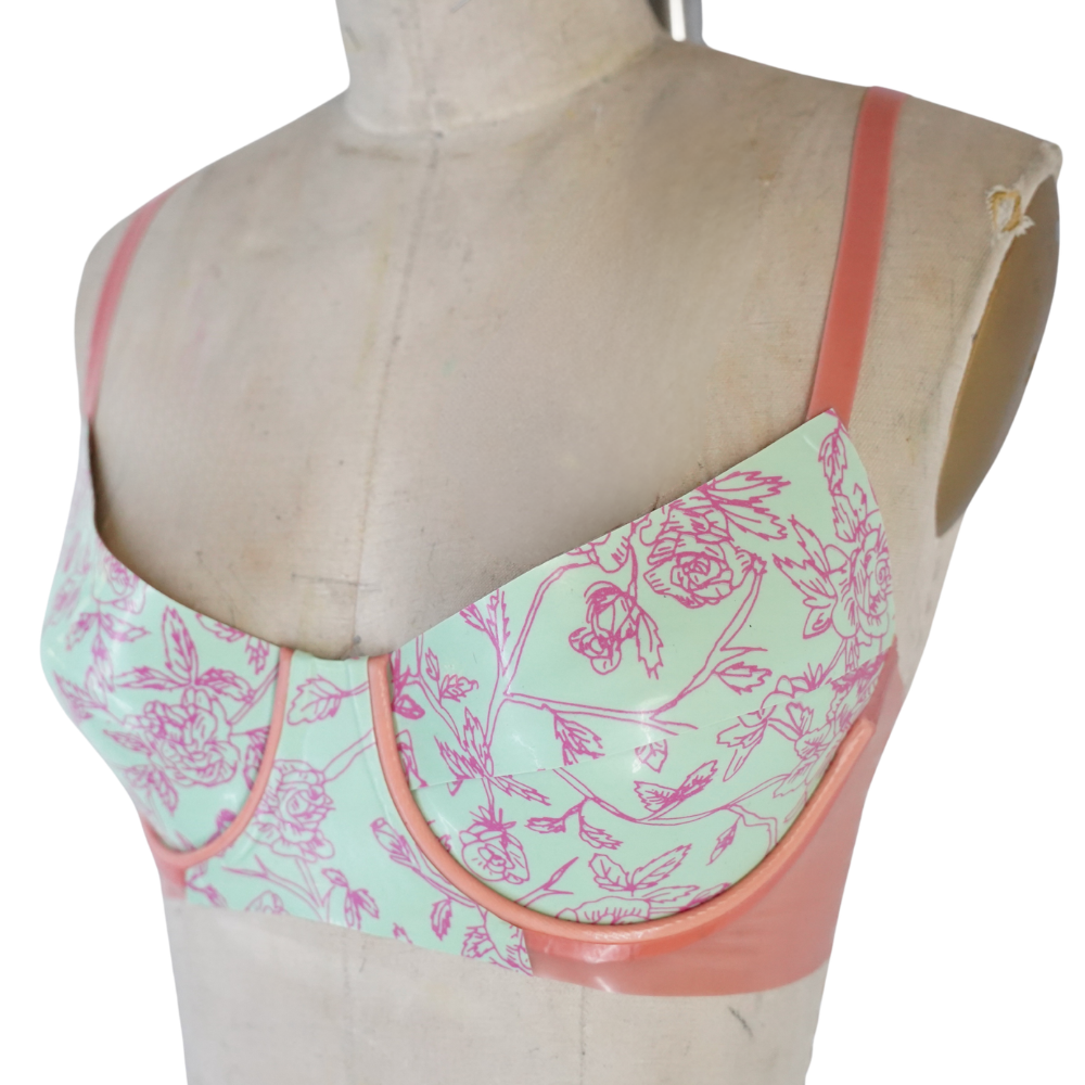A mannequin torso displays a pastel green and pink floral-patterned bra with solid pink straps and trim by Vex Inc. | Latex Clothing. The fitted underwire design is ready to ship, making it a standout in any lingerie collection.