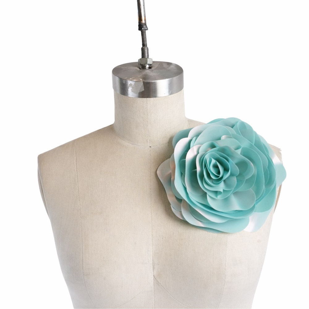 A Vex Latex dress form mannequin showcases a large, hand-crafted teal and white latex statement flower attached to the left shoulder.