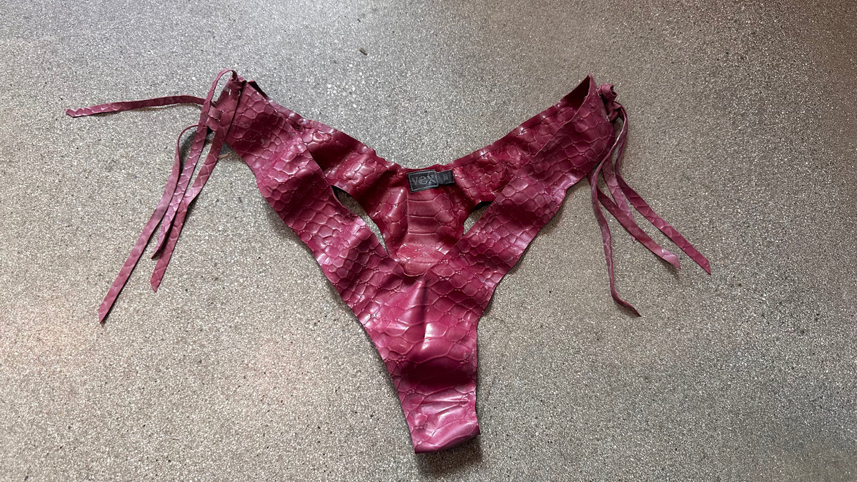 A pair of shiny, textured, dark pink crescent-cut thong-style underwear with a snakeskin pattern and side ties, crafted from natural latex by Vex Latex, is laid flat on a speckled gray surface.