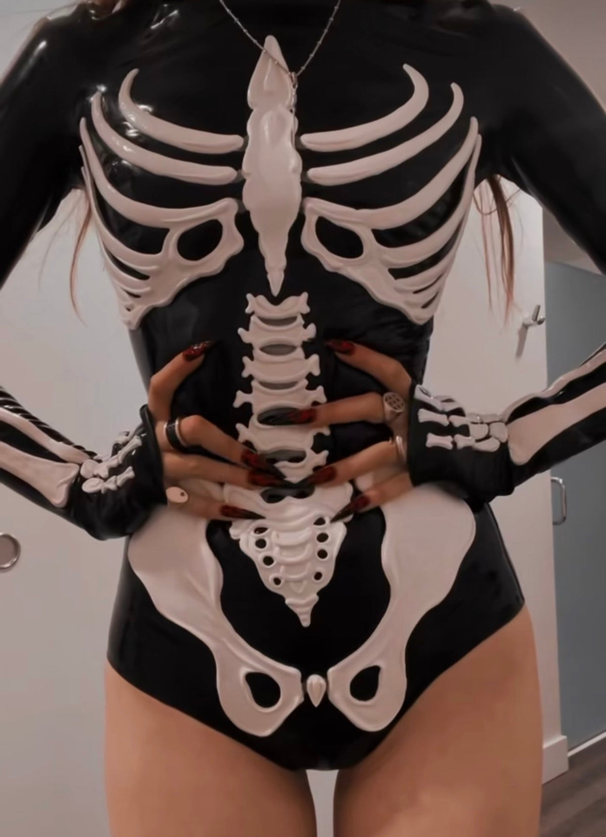 A woman wearing the Vex Skeleton Bodysuit, a black latex bodysuit with sculpted bone-white skeleton bones across the ribs, spine and pelvis, hands resting at her waist. Photographed by @heyitsbunny.