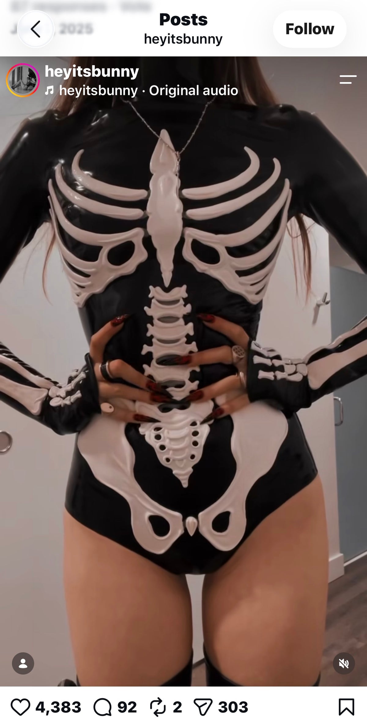 A person wearing a black latex catsuit with a white skeleton design from Vex Clothing, INC stands with their hands on their waist. The background shows part of a door and a white wall.