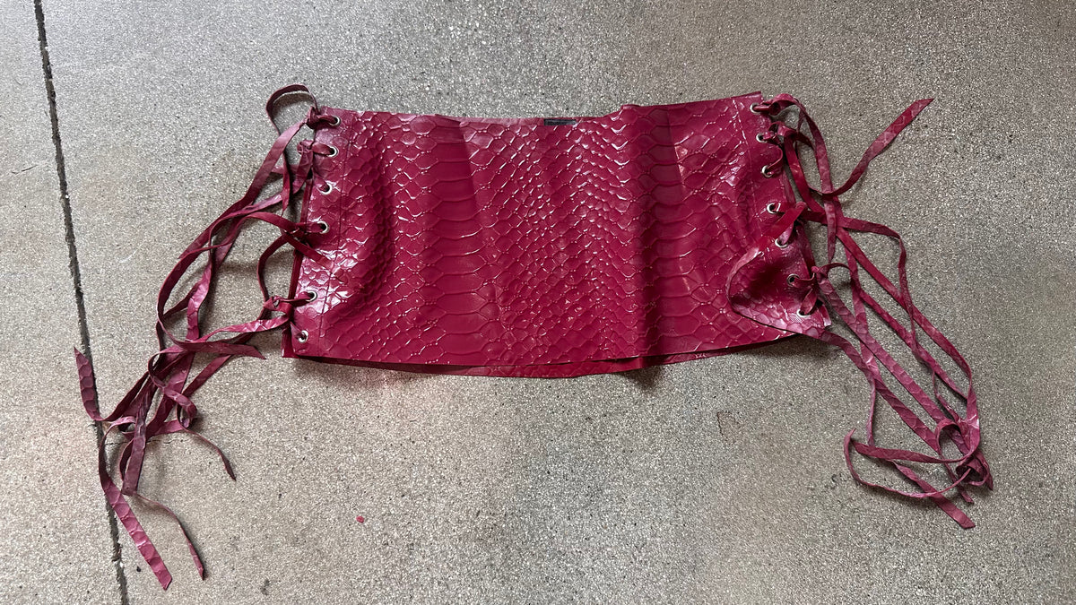 A red faux snakeskin corset from Vex Latex, featuring lace-up sides and long matching ribbons, is laid flat on a gray, speckled floor, perfectly paired with a micro mini latex skirt that highlights its body-skimming silhouette.