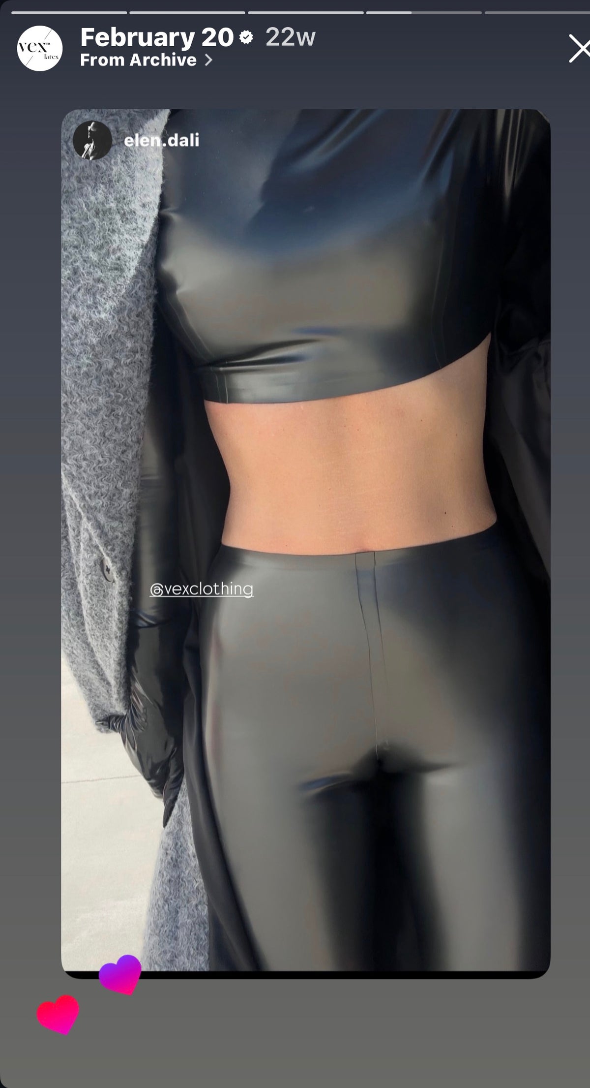 A person is wearing a shiny black crop top and matching high-waisted pants, paired with a gray coat draped over their shoulders—a style evocative of Vex Clothing, INC | Latex Clothing’s iconic high-shine latex designs. The image captures only the torso and upper legs, with two pink heart emojis in the bottom left corner.