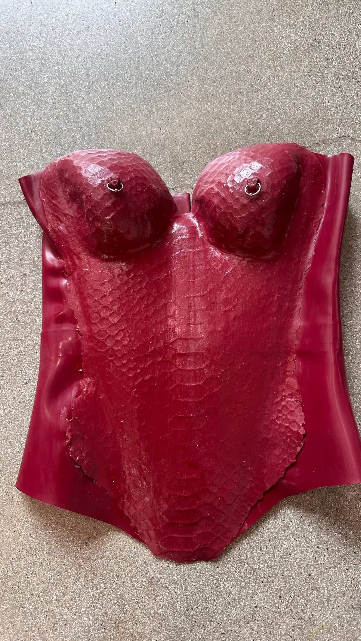 A red, textured molded breastplate by Vex Latex, featuring a scale-like pattern reminiscent of a latex corset and resembling a female torso, is lying flat on a gray, speckled surface.