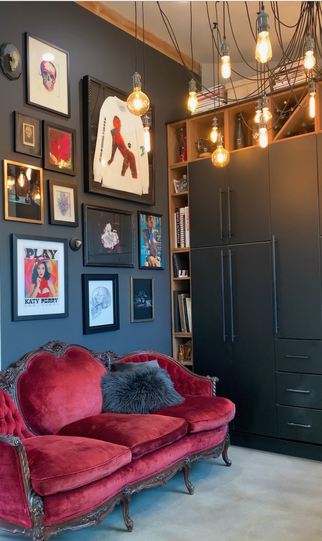 A stylish room with dark walls features a red vintage velvet sofa, framed artworks, hanging exposed lightbulbs, tall black cabinets, bookshelves, and a furry gray pillow on the couch.
