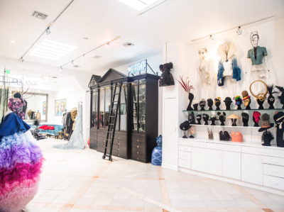 A brightly lit costume shop features colorful, elaborate dresses on mannequins, shelves filled with unique hats and headpieces, and a black ladder leaning against a tall display cabinet.