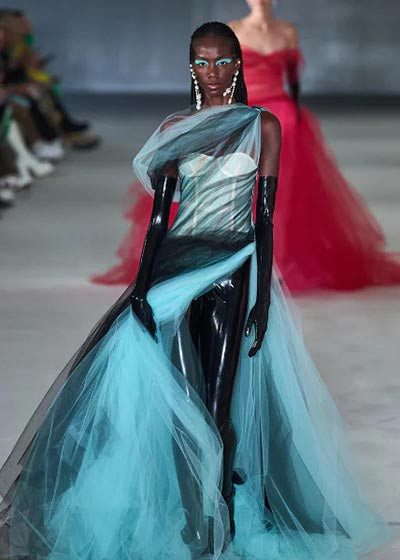 A model walks the runway wearing a sheer blue tulle dress over a black latex bodysuit with long gloves. She has bold blue eye makeup, statement earrings, and slicked-back hair. Another model in a red dress is in the background.