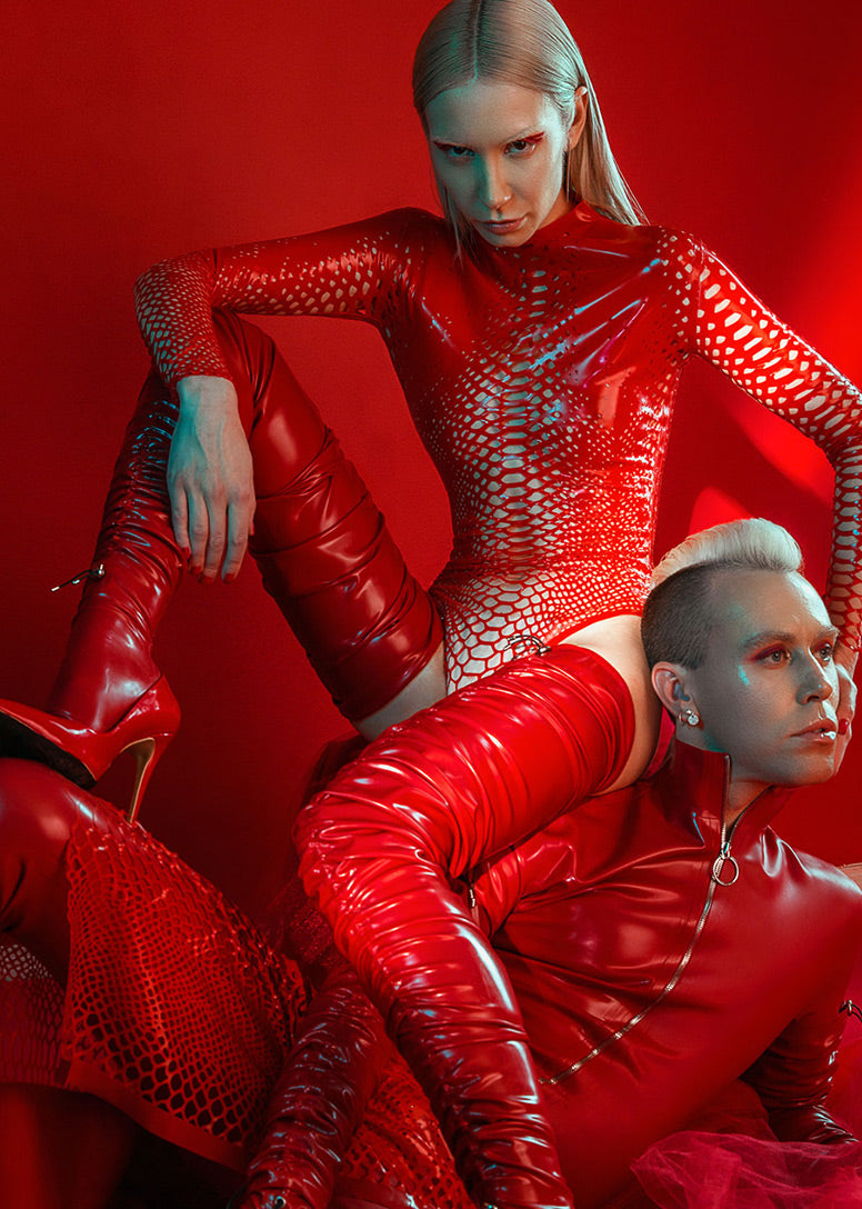 Two models pose against a red background, both wearing shiny, form-fitting red outfits with textured patterns. One model sits confidently above the other, who reclines with one arm raised and intense expressions on both faces.