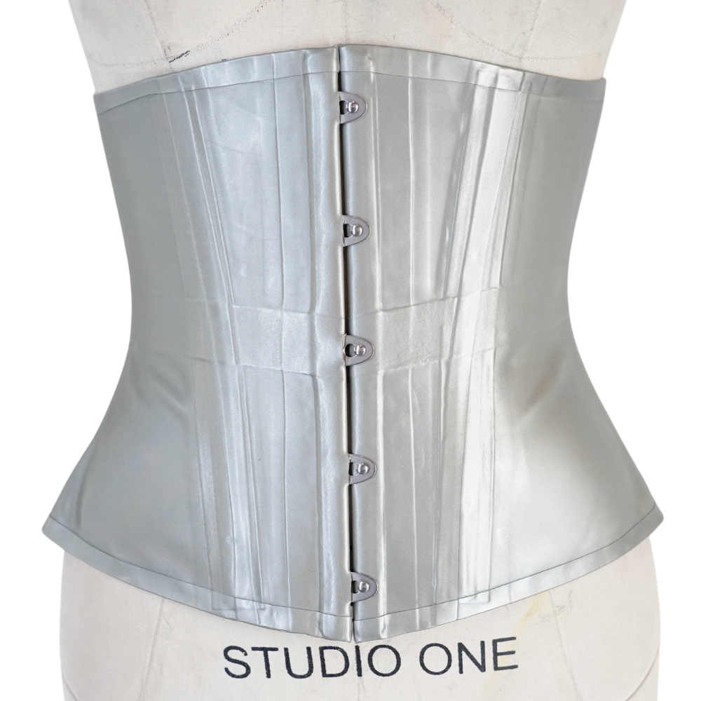 A silver satin corset from Vex Inc. | Latex Clothing is displayed on a STUDIO ONE dress form. Ready to ship, it features steel busk closures, vertical boning for structure, and an hourglass silhouette.
