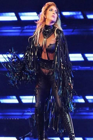 A performer stands confidently on stage under blue lights, wearing a dramatic black outfit with sheer panels, heavy fringe, and thigh-high boots. Her hair is voluminous and styled back, and she looks off to the side.