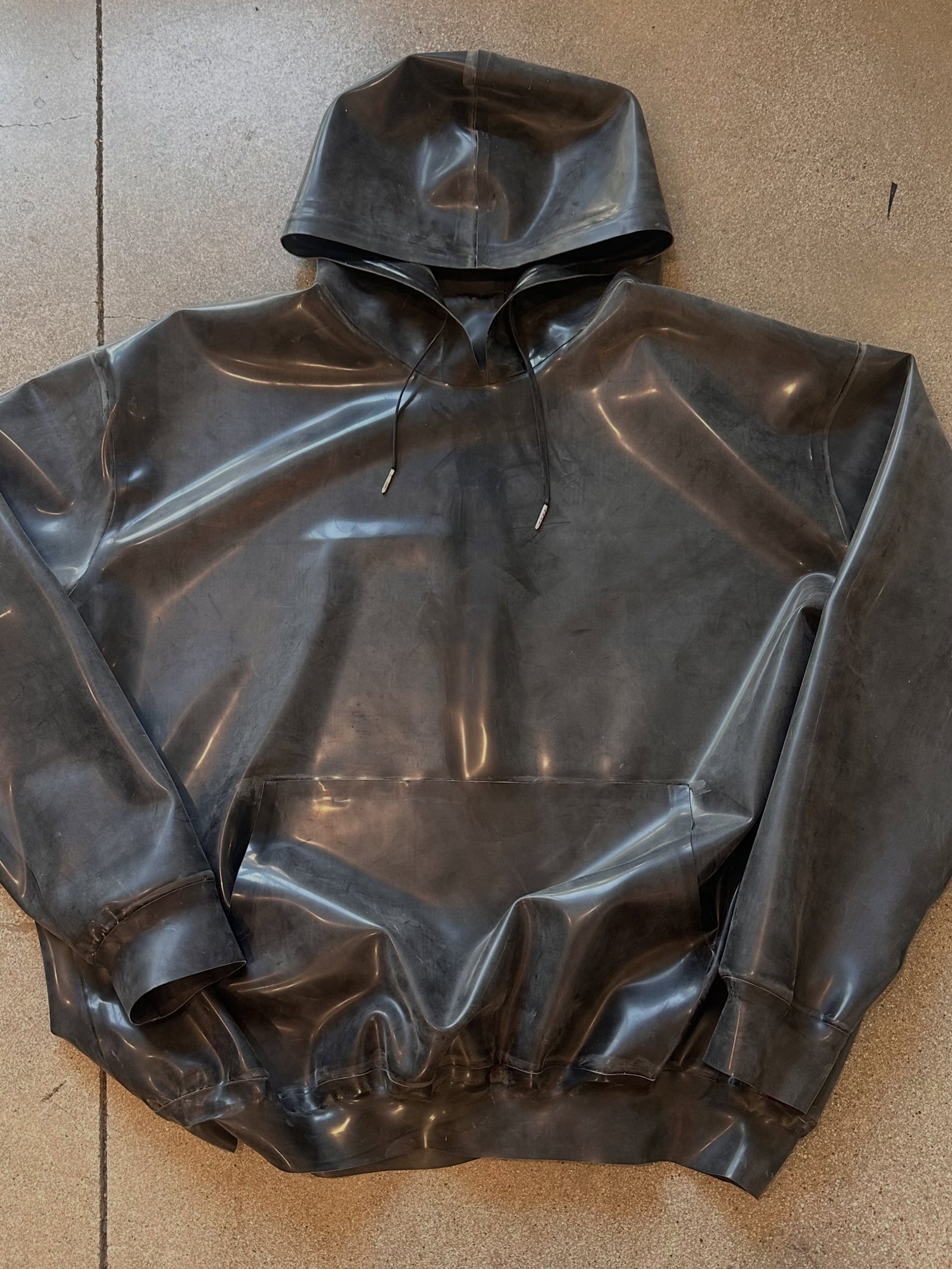 A black, shiny, heavy latex hoodie by Vex Latex, featuring a front pocket and drawstrings, is laid flat on a tan, textured surface.
