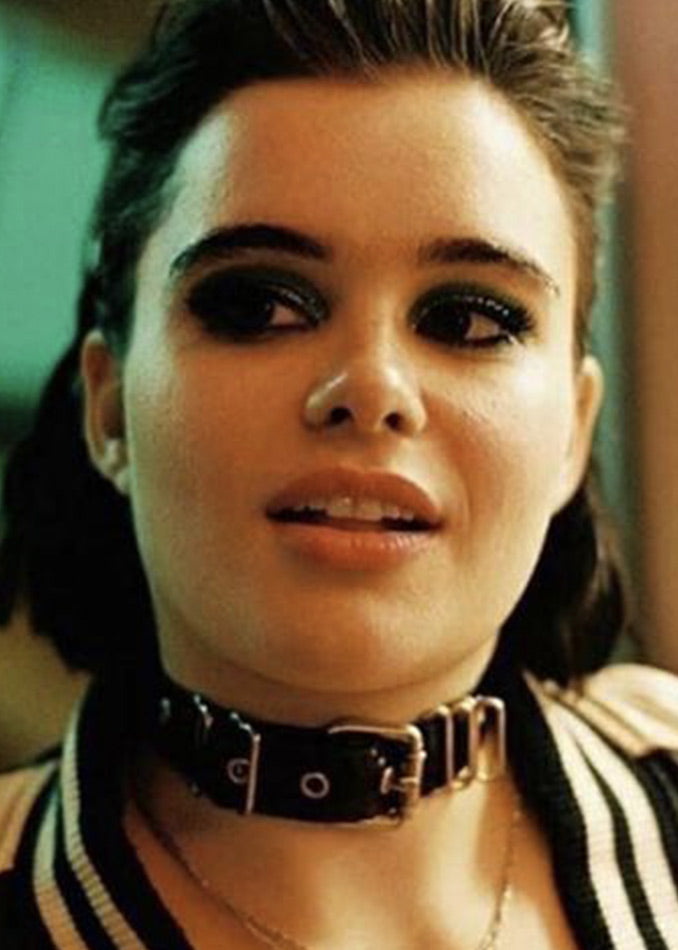 A young woman with dark eye makeup and slicked-back hair wears a thick black choker and a striped jacket, looking slightly to the side with a relaxed expression.