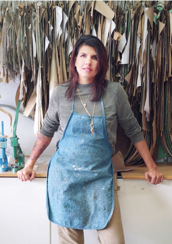 A woman wearing a blue apron and gray shirt stands in front of a wall of hanging paper patterns in a workshop, resting her hands on a worktable and looking at the camera.