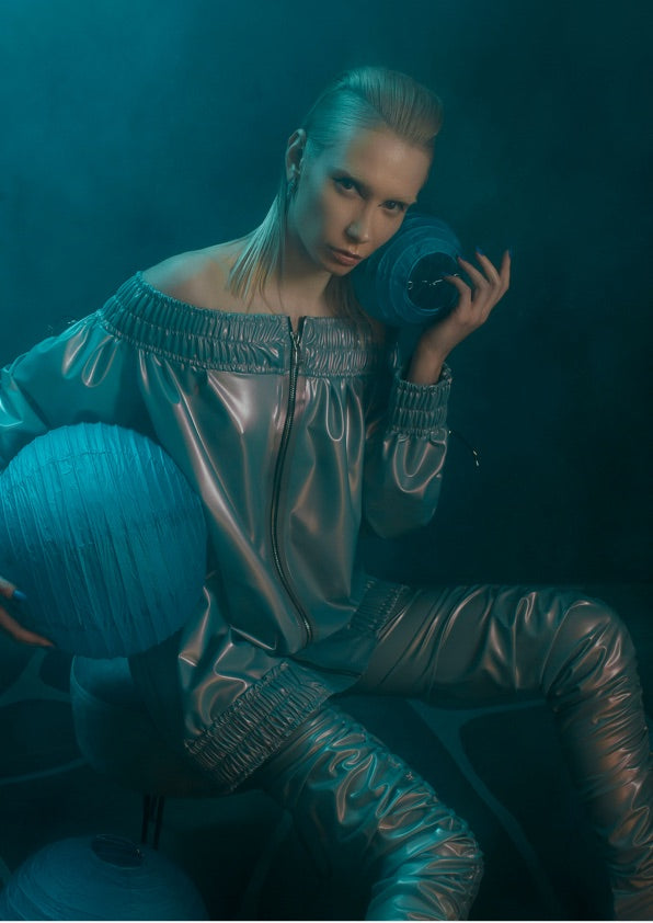 A person wearing a shiny, metallic outfit sits holding blue spherical objects. Their hair is slicked back, and the scene is lit with teal lighting, creating a futuristic, moody atmosphere.